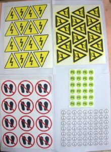 An assortment of polycarbonate membrane overlays and warning labels in various shapes with safety symbols, instructions, and company logos for industrial use.